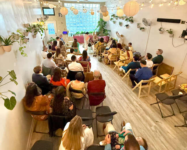 A group of people sit in a brightly lit room adorned with plants and hanging lanterns, facing a speaker at the front.