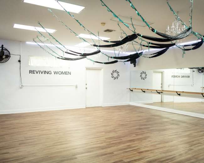The image shows an empty dance studio featuring wooden flooring, large mirrors, and decorated with black fabric drapery and string lights.
