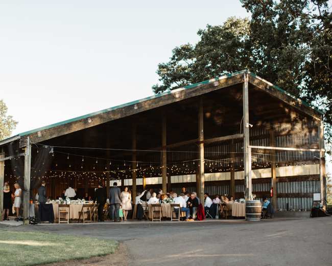 A large outdoor barn setting is filled with tables and guests celebrating, illuminated by string lights.