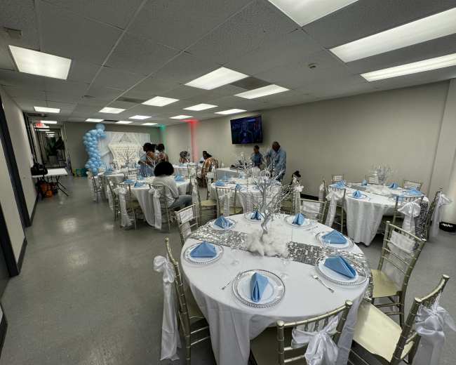 A banquet hall is set up for an event, featuring round tables covered in white tablecloths with blue accents and decorated with elegant centerpieces.