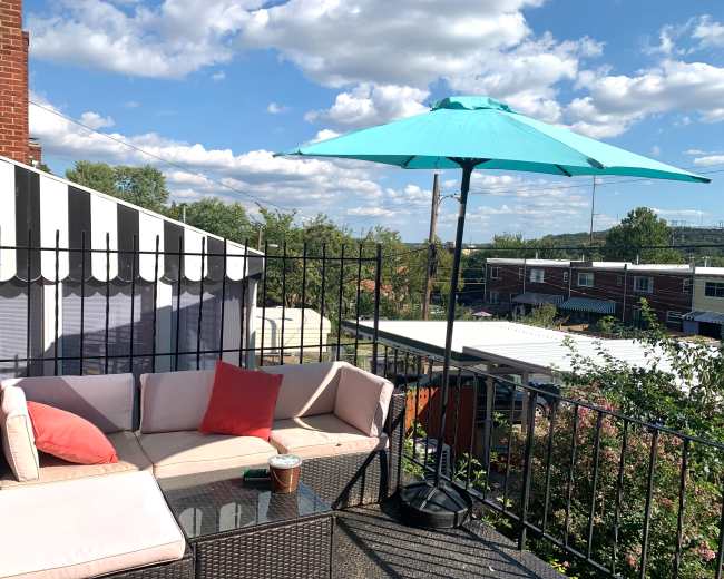 A balcony features a turquoise umbrella over a seating area with a sofa, coffee table, and greenery in the background.