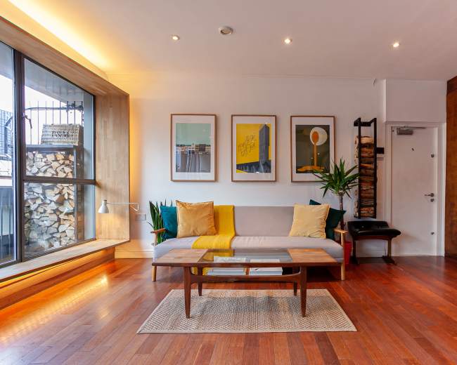The image shows a modern living room with a sofa, a coffee table, and framed artwork on the walls, featuring a large window that allows natural light to enter the space.