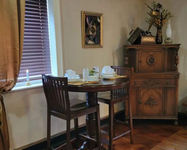 A wooden dining table with two chairs is set for a meal, accompanied by a decorative cabinet and window with blinds.