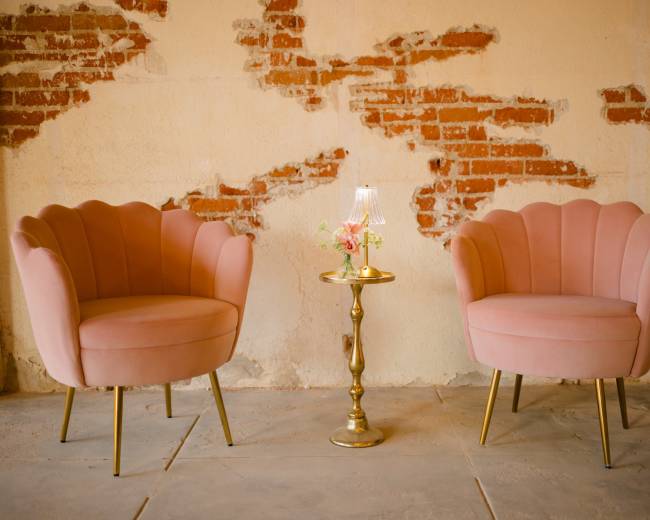 Two pink velvet chairs with scalloped edges are positioned next to a small gold side table, featuring a lamp and a floral arrangement, against a backdrop of exposed brick walls.