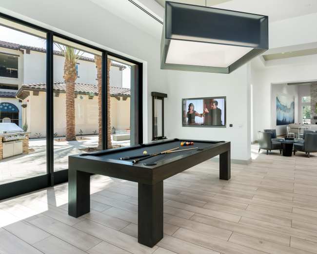 A pool table is positioned in a bright room with large windows, overlooking a courtyard and a seating area with modern furnishings.
