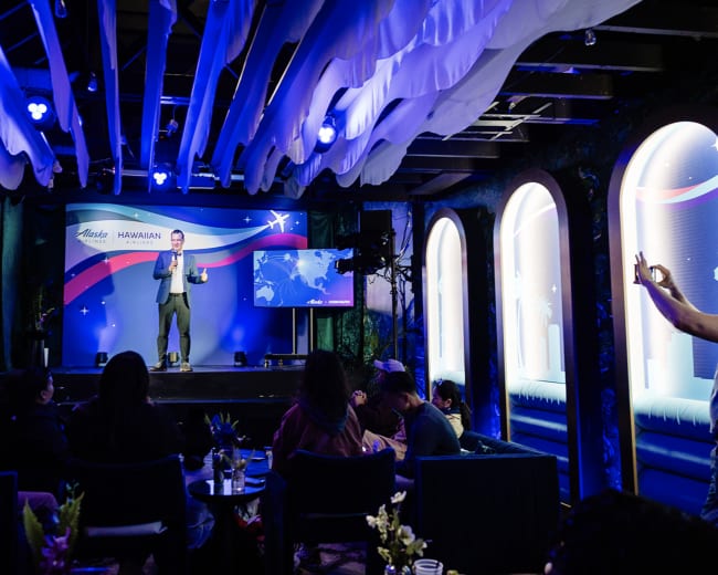 A speaker presents on stage in front of an audience at a Hawaii-themed event in a creatively lit venue.