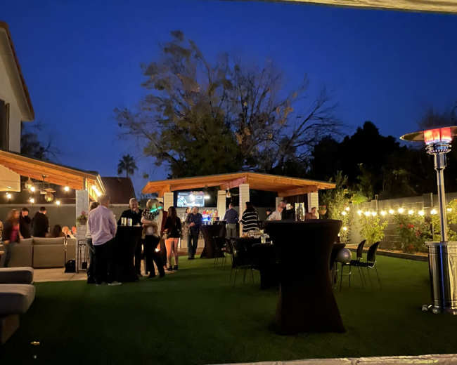 A group of people socializes in an outdoor area with tables and seating under twilight, surrounded by trees and illuminated by string lights.