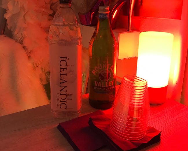A plastic bottle of Icelandic glacial water and a glass bottle of Mountain Valley sparkling water sit next to a stack of red plastic cups on a wooden surface, illuminated by a red lamp.