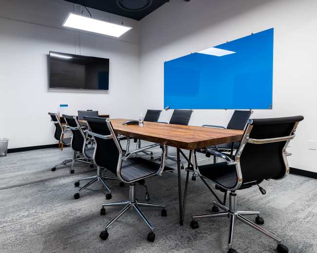 The image shows a modern conference room featuring a wooden table surrounded by black chairs, with a television on the wall and blue whiteboards.