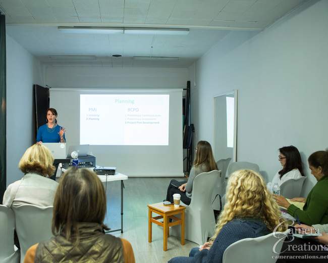 A woman delivers a presentation in a bright room, facing an audience seated in chairs, with a projector displaying slides on a screen.