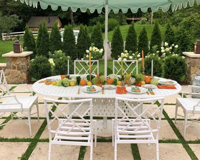 A round outdoor dining table is set for a meal under a mint-green umbrella, surrounded by white chairs and lush greenery.
