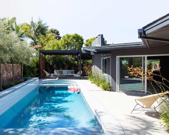 A modern backyard featuring a swimming pool with a lounger and a shaded seating area surrounded by greenery.