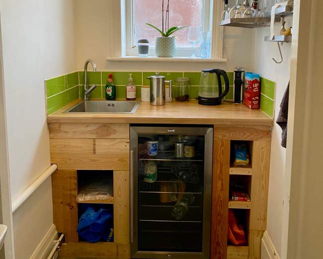 The image shows a small kitchen area featuring a wooden cabinet with a sink, a kettle, and a compact refrigerator, along with green tiled backsplash and a window above with a potted plant.