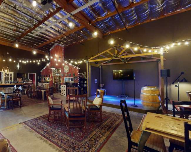 The image shows an indoor space with wooden furniture, string lights, and a stage area, resembling a rustic gathering venue.