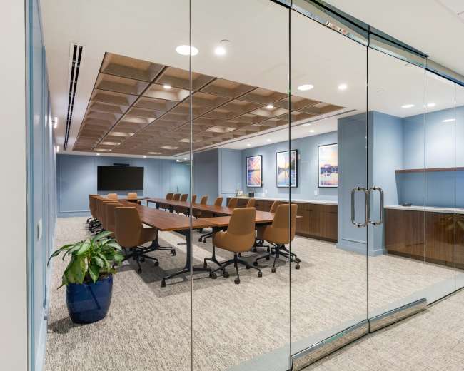 The image shows a modern conference room with a large table, ergonomic chairs, and wall-mounted artwork, enclosed by glass walls.