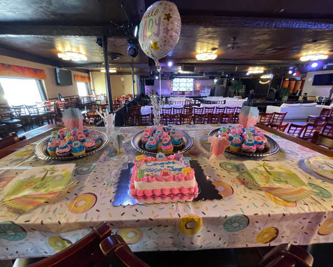 A decorated table set up for a celebration, featuring multiple colorful cakes, cupcakes, and themed tableware in a venue with dim lighting.