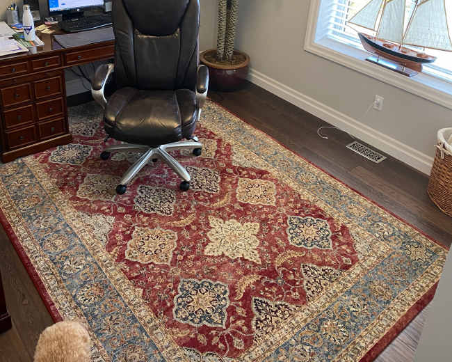 An office space features a desk with a computer, a swivel chair, a decorative palm plant, and a model sailboat near a window, all set on a patterned area rug.
