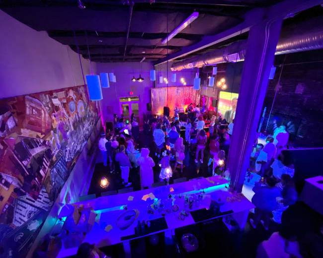 A crowded indoor venue features vibrant lighting and a large mural on the wall, with people socializing and enjoying the atmosphere.
