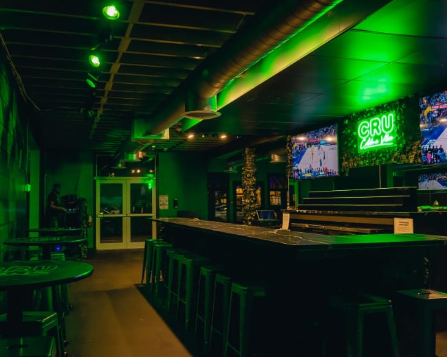 The image shows a dimly lit bar with green accents, featuring a long counter, high stools, and screens displaying sports events.