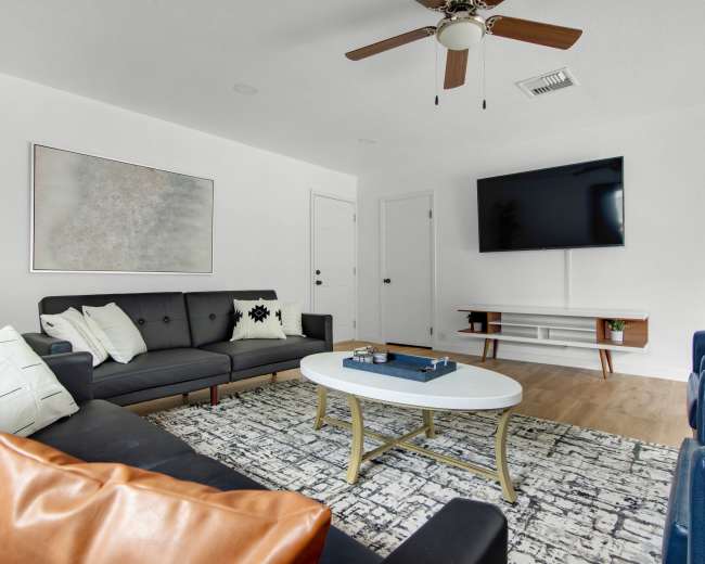A modern living room features a dark sectional sofa, a central coffee table, and a wall-mounted television, with wooden flooring and a light color scheme.