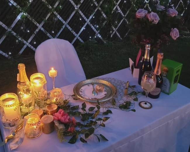 A decorated table for two is set with candles, champagne bottles, and flowers, against a backdrop of illuminated greenery.