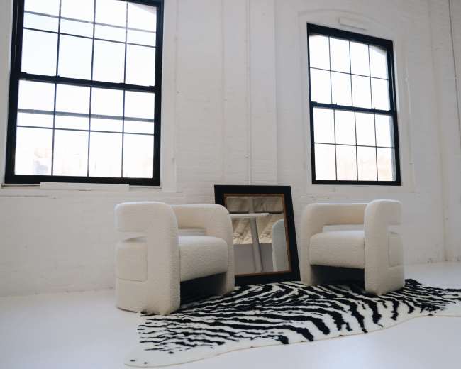 Two white upholstered chairs are positioned on a zebra print rug in a bright room with large windows and a mirror leaning against the wall.