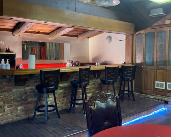A bar area with a wooden counter and high black stools, set against a backdrop of exposed brick walls and wooden beams.