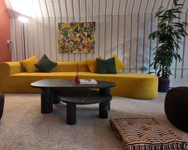 The image shows a modern living space featuring a yellow curved sofa, a black marble coffee table, and a large abstract painting on the wall, along with a plant and comfortable seating.