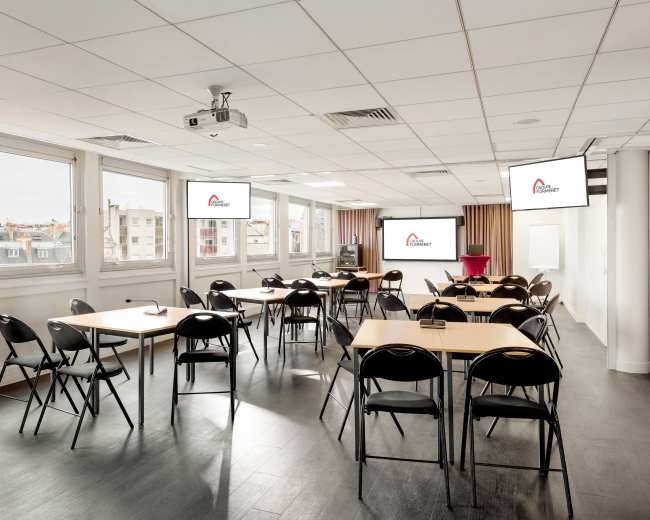 A modern classroom setup features several tables and chairs arranged for a presentation, with large screens displaying a logo on the walls.
