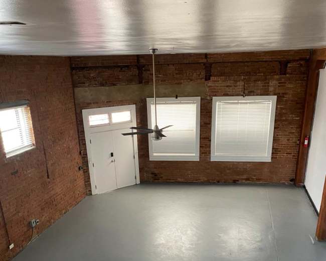 The image shows a spacious room with exposed brick walls, large windows, and a gray concrete floor.