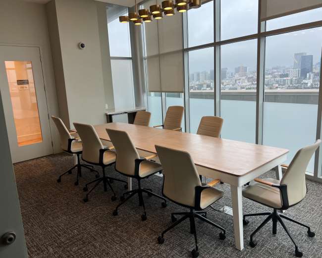A conference room features a long wooden table surrounded by six beige rolling chairs, with large windows showcasing a city skyline in the background.