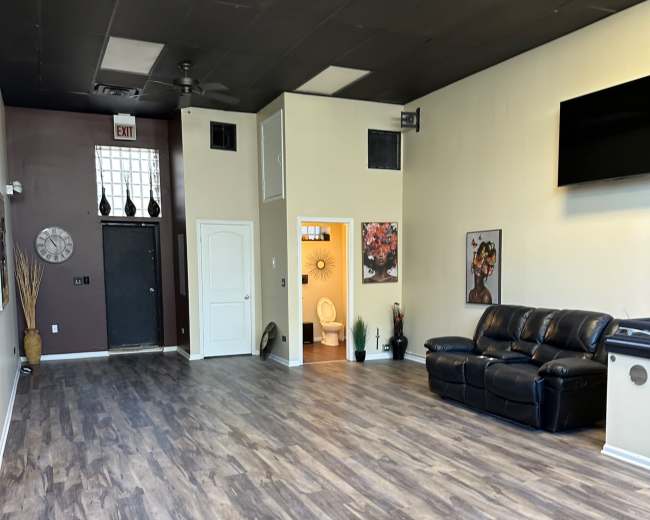 The image shows an empty room with wooden flooring, a black leather couch, a wall-mounted television, and a doorway leading to a bathroom area.