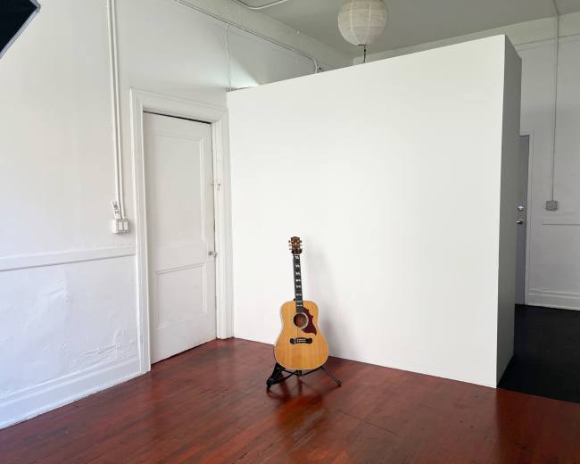 A wooden guitar stands on a stand in a sparsely furnished room with a white wall and a door.