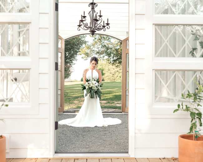 A bride in a white gown holds a bouquet while standing in a doorway with large wooden doors and a chandelier overhead.