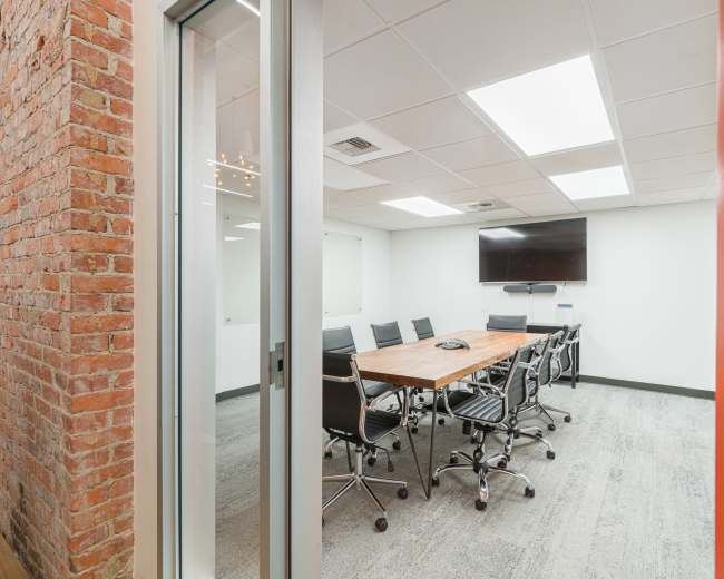 The image shows a modern conference room featuring a large wooden table surrounded by black office chairs, a wall-mounted television, and exposed brick on one side.
