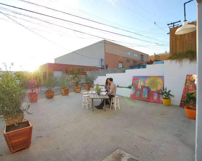 A woman sits at a table in a spacious outdoor area surrounded by potted plants and colorful murals.
