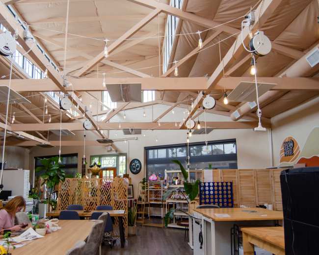 The image shows a bright, open workspace with wooden beams, overhead lighting, and various furniture arrangements for different tasks.