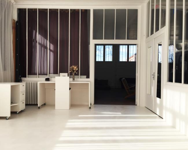 The image shows a bright, minimalistic office space with large windows, light-colored furniture, and a clean floor.