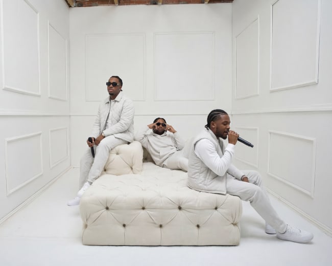 Three men dressed in white are performing and posing on a white tufted sofa in a minimalist, all-white room.