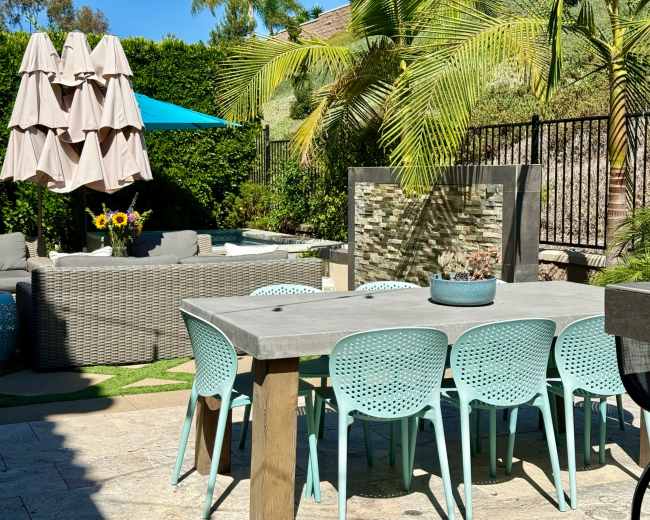 A large outdoor patio features a dining table surrounded by turquoise chairs, a style of furniture, a fountain, and a sunshade umbrella.