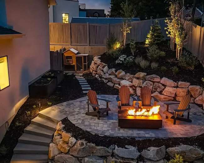 The image shows a backyard patio featuring a fire pit surrounded by four wooden chairs, stone landscaping, and a small shed in the background.