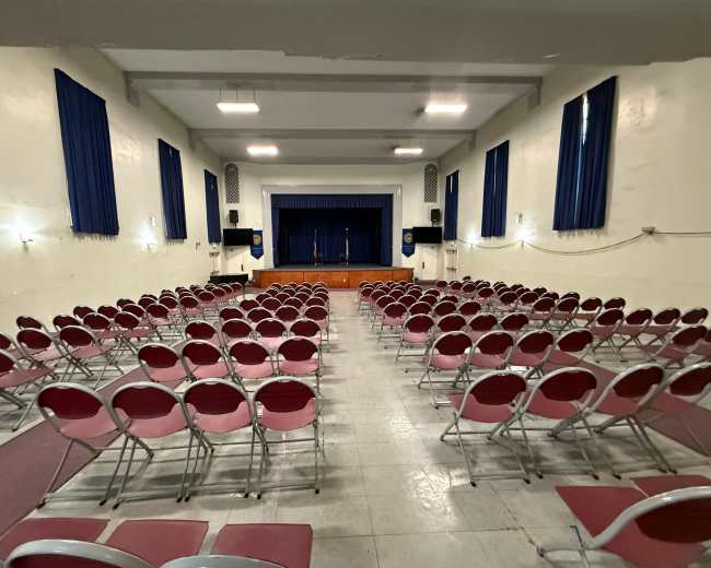 The image shows a large auditorium with rows of red-cushioned chairs arranged facing a stage at the front.