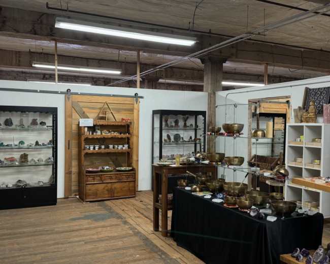 The image shows a retail space featuring glass display cases filled with crystals and stones, wooden shelves, and several tables showcasing various items for sale.