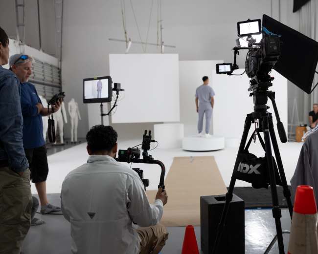 A film crew sets up equipment in a white studio, with a model on a circular platform in the background.