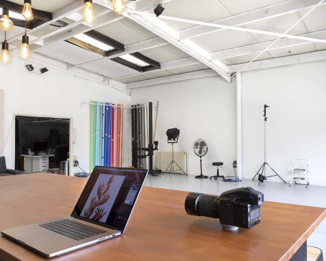A spacious photography studio features a wooden table with a laptop and camera, surrounded by studio lighting equipment and backdrops.