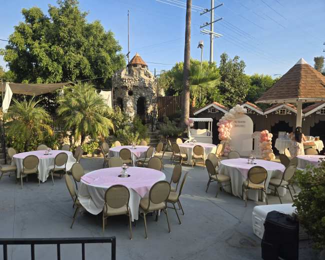 The image shows an outdoor event space with round tables covered in pink tablecloths, surrounded by greenery and a stone structure in the background.