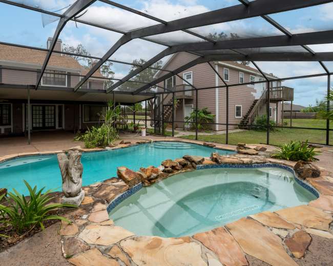 The image shows a spacious outdoor area with a swimming pool and a hot tub, surrounded by a screened enclosure and a two-story house in the background.