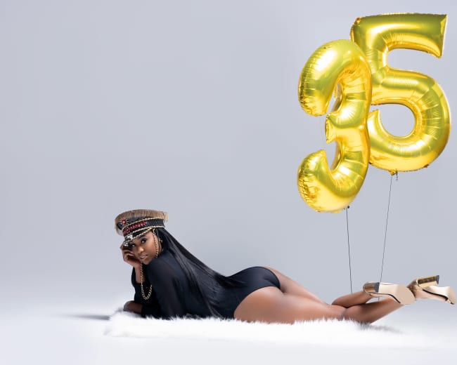 A woman in a black outfit poses on a white fur rug with large gold balloons shaped like the number 35 behind her.