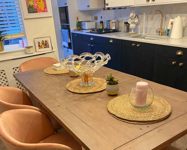 A wooden dining table is set with round placemats, decorative glassware, and chairs with pink upholstery in a modern kitchen.