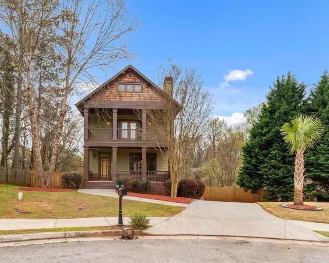 The image shows a two-story house with a wooden exterior, situated at the end of a driveway, surrounded by trees and shrubs.